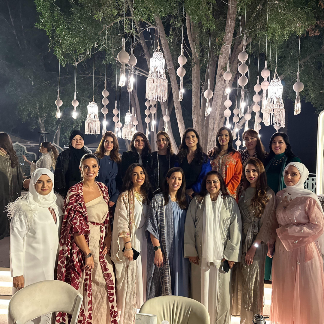 A group of women posing together at a nighttime event under hanging decorative lights and trees, wearing traditional and formal garments in white, beige, pink, blue, and patterned fabrics.