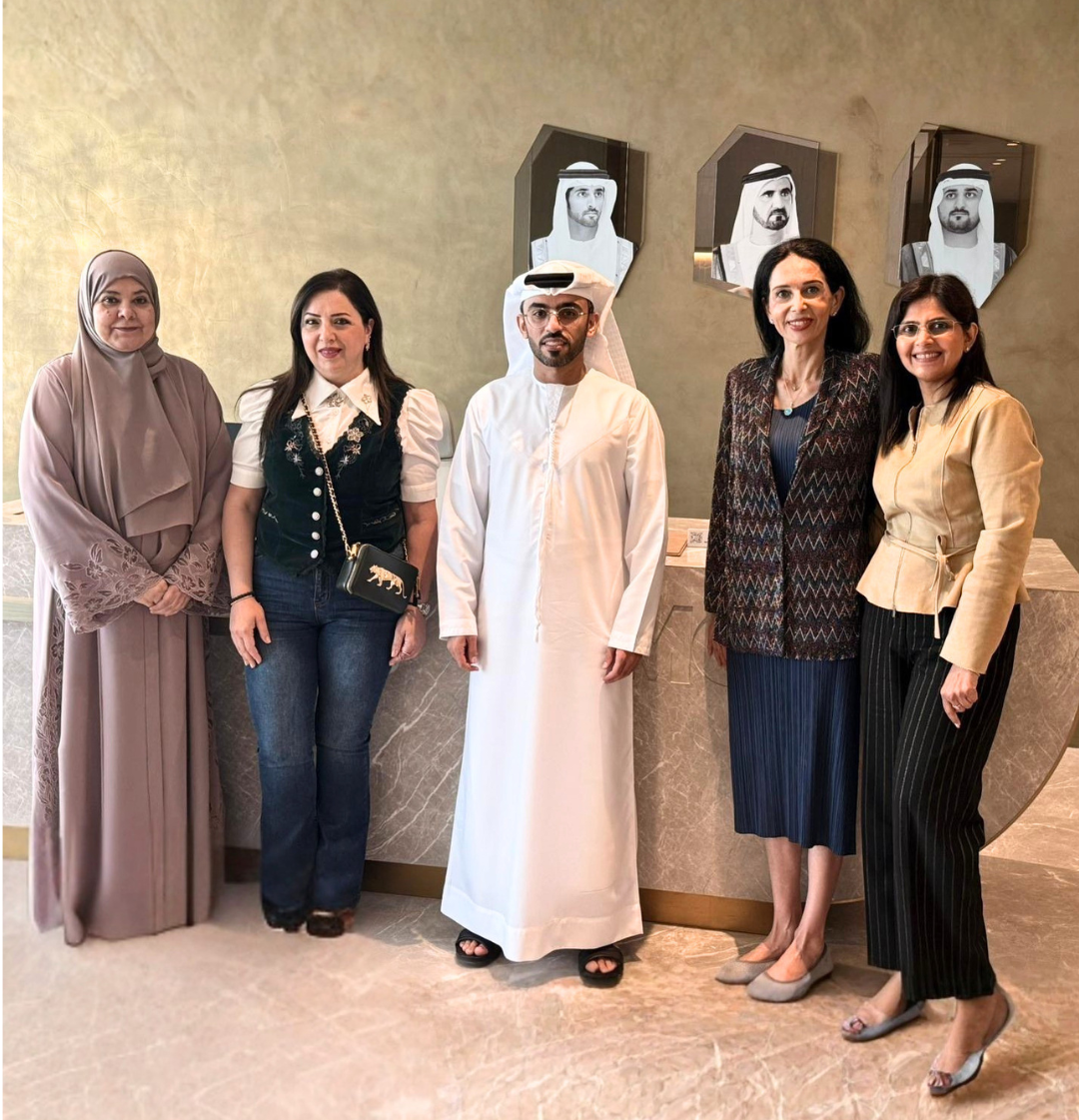 Five people posing together in front of a beige wall with framed portraits. The group includes a woman in a taupe hijab and dress, a woman in a dark vest and jeans, a man in a white thobe and head covering, a woman in a patterned jacket and blue skirt, and a woman in a beige jacket and black pants.